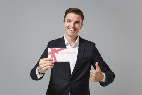 Smiling young business man in classic black suit shirt posing isolated on grey wall background. Achievement career wealth business concept. Mock up copy space. Hold gift certificate showing thumb up.