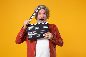 Shocked elderly gray-haired mustache bearded man in red leather jacket isolated on yellow orange background. People lifestyle concept. Mock up copy space. Hold classic black film making clapperboard.