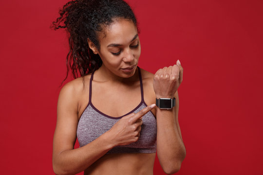 Beautiful young african american sports fitness woman in sportswear working out isolated on red background. Sport exercises healthy lifestyle concept. Pointing finger on smart watch with empty screen.