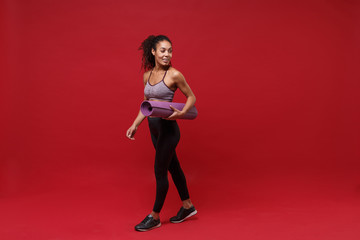 Fototapeta na wymiar Smiling young african american sports fitness woman in sportswear working out isolated on red background studio portrait. Sport exercises healthy lifestyle concept. Hold fitness mat, looking aside.