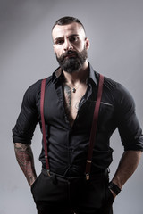 Ragazzo barbuto con Tatuaggi in tutto il corpo, vestito con camicia nera e bretelle rosse, isolato...