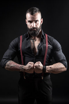 Ragazzo barbuto con Tatuaggi in tutto il corpo,  con pugni chiusi, vestito con camicia nera e bretelle rosse, isolato su sfondo nero.