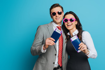happy businessman and businesswoman in sunglasses holding passports and air tickets on blue background
