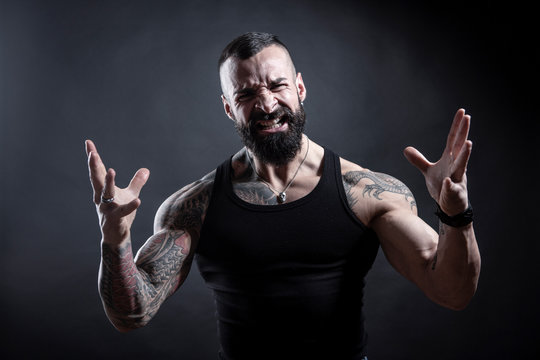 Uomo con barba compeltamente tatuato in canottiera guarda molto irritato con , le braccia e le mani in movimento , isolato su sfondo nero