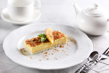 Desserts. Fruit cake with sponge cake and yellow mousse. Mint, yellow slice and powdered sugar on a light plate. Cup6 teapot near plate. Copy space