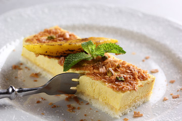 Desserts. Fruit cake with sponge cake and yellow mousse with fork. Mint, yellow slice and powdered sugar on a light plate. Copy space