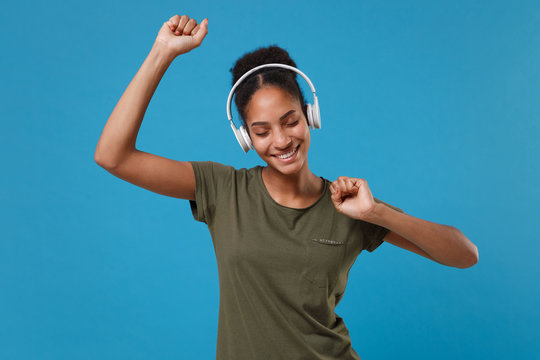 Cheerful young african american woman girl in casual t-shirt posing isolated on blue background. People lifestyle concept. Mock up copy space. Listen music with headphones dancing keeping eyes closed.