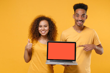 Smiling friends couple african american guy girl in casual clothes isolated on yellow background. People lifestyle concept. Point finger on laptop pc computer with blank empty screen showing thumb up.