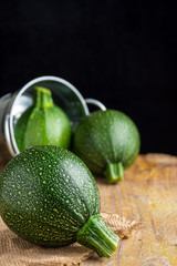 Close-up of a round zucchini lying on burlap cloth, two more zucchini in the background in a metal bucket, with selective focus, on rustic table and black background, in vertical