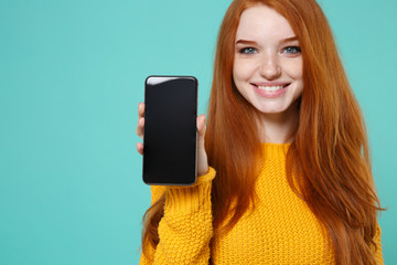 Cropped image of smiling young redhead girl in yellow sweater posing isolated on blue turquoise background. People lifestyle concept. Mock up copy space. Hold mobile phone with blank empty screen.