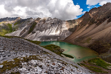 Naklejka premium A small lake among the mountains. White and brown mountain nearby. Stone scree in the foreground. White clouds on a blue sky. Horizontal.