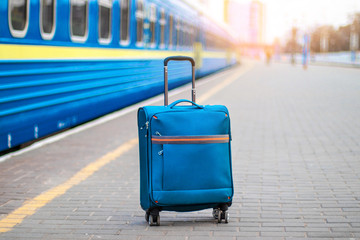Blue carry on suitcase with wheels and a metal handle stands on the peron of the station. Passenger...