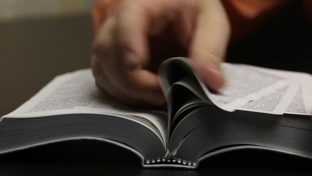 On the table lies an open Bible. The man quickly turns the pages in search of the desired chapter. Finds and affirmatively pokes a finger in the right place. Close-up shot.