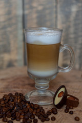 Latte coffee / cappuccino in a transparent glass with a pen on a wooden background. Coffee beans. Chocolate candies with nuts. Morning coffee concept.