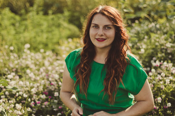 Young curly Caucasian girl in a green dress looks at the camera and smiles in a flower field on a sunny day. Spring or summer panorama photo