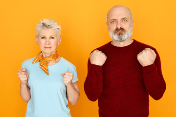 Studio image of elderly couple standing next to each other expressing negative emotions, being angry with high prices or low pension payments, clenching fists, having mad furious facial expression