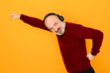 Studio image od energetic active bearded European male pensioner using bluetooth wireless earbuds...