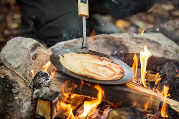 Man and woman making pancakes on campfire in forest on shore of lake, making a fire, grilling. Happy couple exploring Finland. Scandinavian landscape. 