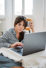 Working young brunette woman freelancer in shirt with laptop and cup of tea work at home in the bright interior