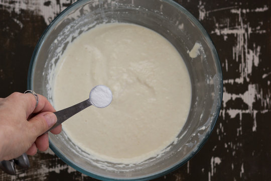 Top view of woman hand putting tablespoon of soda into swollen semolina as ingredient for baking mannik caressole on the brown background
