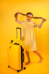 Portrait of a beautiful little girl in a yellow dress with pigtails standing near a suitcase. positive girl joyfully looks at the camera holds hair in her hands