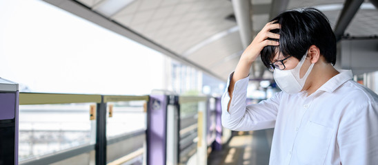 Fototapeta premium Asian man wearing face mask feeling headache at skytrain station platform. Coronavirus (COVID-19) outbreak prevention in public transportation. Health awareness for PM2.5 air pollution protection.