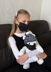 A girl with blonde hair dressed in a black and white school uniform hugs a soft toy bear. Both are in black medical masks. School is under quarantine due to the pandemic of Covid-19. Home schooling