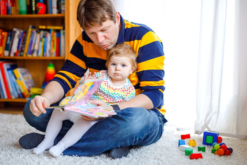 Young father reading book with his cute adorable baby daughter girl. Smiling beautiful child and man sitting together in living room at home. Toddler hearing to dad.