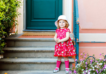 Portrait of beautiful little gorgeus lovely toddler girl in pink summer look clothes, fashion dress, knee socks and hat. Happy healthy baby child posing infront of colorful house.