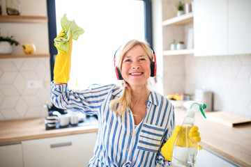Portrait of senior woman with headphones and gloves cleaning indoors at home.