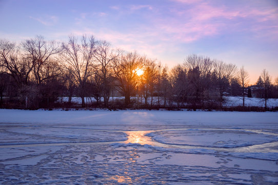 Evening sun overlooking a frozen pond lit with pink sunset sun rays through bare trees branches in Unionville, Markham, Ontario, Canada
