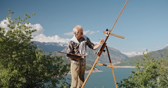 Senior caucasian man is standing in front of easel, drawing a picture. Experienced artist making an artpiece, inspired by landscape - retirement, inspiration concept 4k