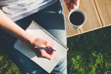 young woman's hands writing notes on notebook with pen on her knees outdoor and drinking coffee...