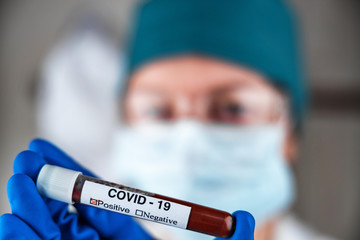 A nurse in a respiratory mask holds a test tube labeled COVID 19, with a positive blood test for a new rapidly spreading coronavirus, close-up, shallow depth of field, selective focus. The concept of