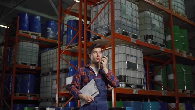 Panning waist up shot of young material handler in uniform talking on walkie-talkie standing by racks with steel barrels and containers in factory storage