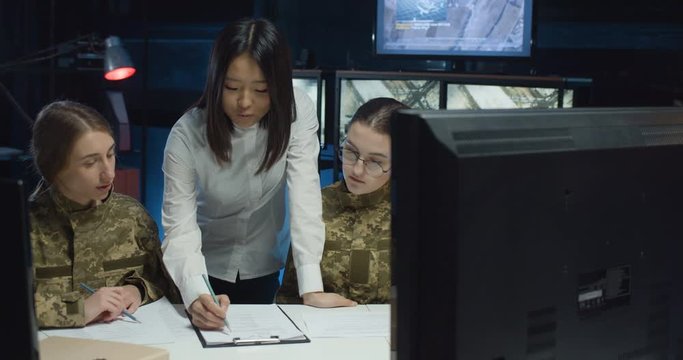 Two young Caucasian girls in uniforms of soldiers studying in military controlling office. Asian female teacher teaching women of army to be dispatchers in monitoring room.