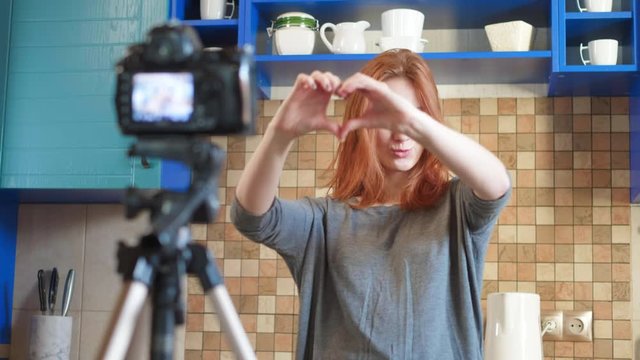 Food blogger girl influencer is recording a video or podcast in the kitchen. Makes lifestyle blog vlog, shows a heart sign from hands. A woman communicates with subscribers, asks to like the video
