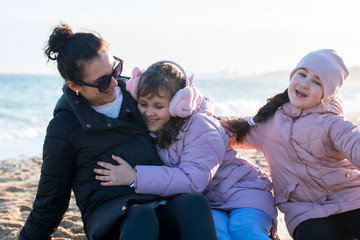 a young woman and two little girls in jackets and earmuffs play and hugs on the beach