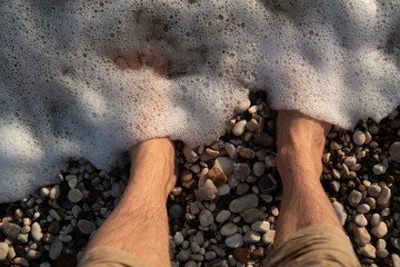 Sea foam washes men's feet standing on a pebble beach