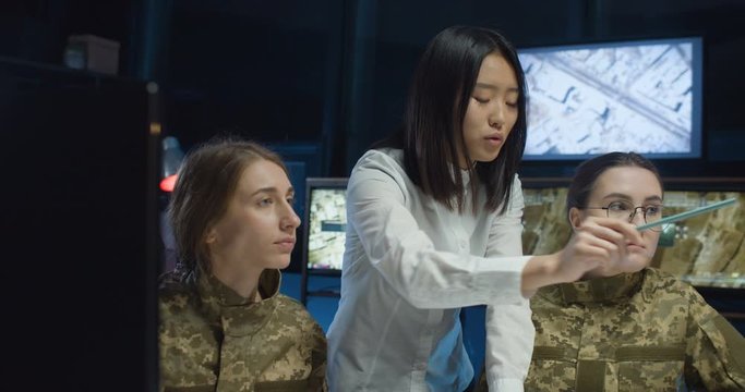 Asian female educator teaching Caucasian girls of army studies in monitoring room and showing something in dark office with screens. Teacher with students in military university