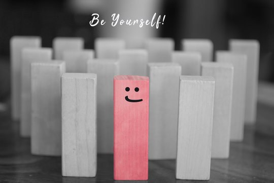 Inspirational Motivational Quote - Be Yourself. With Background Of Wooden Blocks In Same White Color And One Wood Block In Pink Red Color, With Cute Funny Smiling Face Emoticon On Wooden Block.