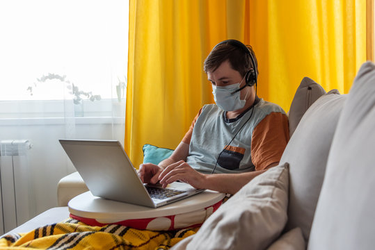 A young sick man in a mask sits on a sofa with a laptop and a headphones and works remotely. The concept of home quarantine and self-isolation.