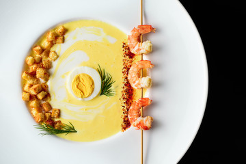 Warm cream soup with cheese, egg, green dill, shrimp and spices, fried croutons, in a white round plate on a black background