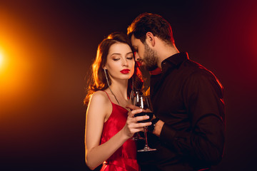 Handsome man holding glass of red wine near elegant girlfriend on black background with lighting