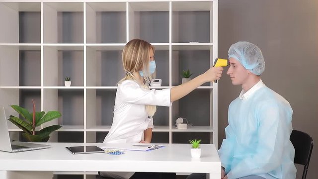 Doctor in a white coat and a medical mask measures the temperature of a patient in the medical office with an infrared non-contact thermometer.