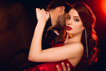 Naklejka premium Elegant girl in red dress looking at camera while hugging boyfriend on black background with lighting