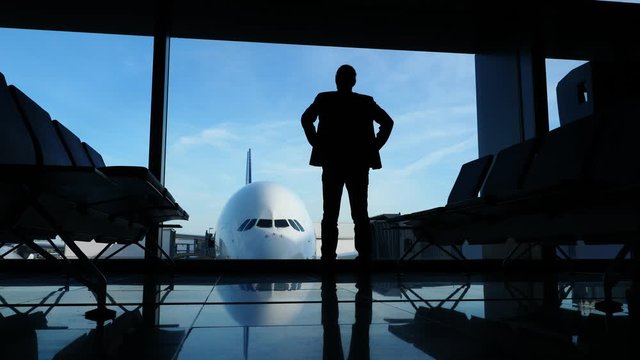 Silhouette of man in suit standing with arms on hips against terminal window. Passenger look out to large airliner parked at gate apron area. Low camera, dark economy class lounge interior