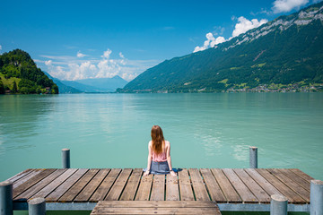 Back view of a young girl sitting on a pier. Beautiful turqouise lake, mountains and sky background. Vacation and traveling concept