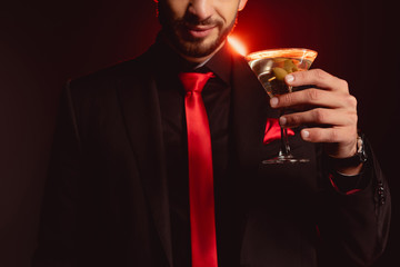 Cropped view of elegant man holding glass of cocktail on black background with lighting