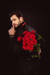 Side view of handsome man holding bouquet of roses and showing secret sign isolated on black